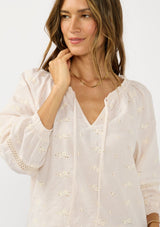 [Color: Tan/Natural] A detail image of a brunette model wearing a beige checkered blouse with floral embroidery, lace details, and a split v neckline with ties. 