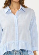 [Color: Light Blue] A detail image of a brunette model wearing a blue striped blouse with a button front, collared neck, and a high low hemline with ruffle trim.