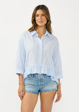 [Color: Light Blue] A front facing image of a brunette model wearing a blue striped blouse with a button front, collared neck, and a high low hemline with ruffle trim.