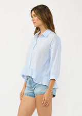 [Color: Light Blue] A side facing image of a brunette model wearing a blue striped blouse with a button front, collared neck, and a high low hemline with ruffle trim.