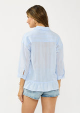 [Color: Light Blue] A back facing image of a brunette model wearing a blue striped blouse with a button front, collared neck, and a high low hemline with ruffle trim.