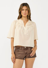 [Color: Natural] A front facing image of a brunette model wearing a beige cotton blouse with a ruffled yoke, half sleeves, and a button front.