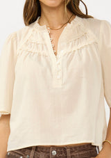 [Color: Natural] A detail image of a brunette model wearing a beige cotton blouse with a ruffled yoke, half sleeves, and a button front.