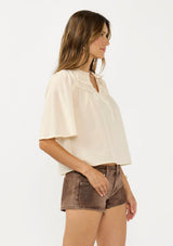 [Color: Natural] A side facing image of a brunette model wearing a beige cotton blouse with a ruffled yoke, half sleeves, and a button front.