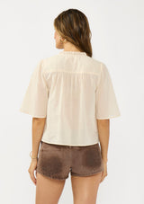 [Color: Natural] A back facing image of a brunette model wearing a beige cotton blouse with a ruffled yoke, half sleeves, and a button front.