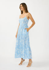 [Color: Blue/Ivory] A side facing image of a brunette model wearing a blue and white floral maxi dress with a scoop neckline, adjustable spaghetti straps, button front, and side pockets. 