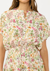 [Color: Blush/Rose] A detail image of a brunette model wearing a pink and green floral print mini dress with short dolman sleeves, a button front, a smocked waist, and a ruffle tiered hem with layers. 