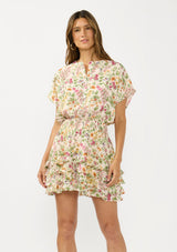 [Color: Blush/Rose] A front facing image of a brunette model wearing a pink and green floral print mini dress with short dolman sleeves, a button front, a smocked waist, and a ruffle tiered hem with layers. 