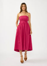 [Color: Magenta] A front facing image of a brunette model wearing a pink strapless midi dress with a gathered bust, side pockets, and a smocked back.