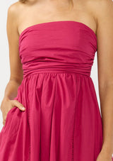 [Color: Magenta] A detail image of a brunette model wearing a pink strapless midi dress with a gathered bust, side pockets, and a smocked back.