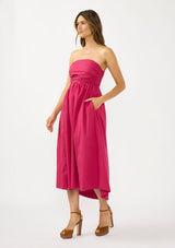 [Color: Magenta] A side facing image of a brunette model wearing a pink strapless midi dress with a gathered bust, side pockets, and a smocked back.