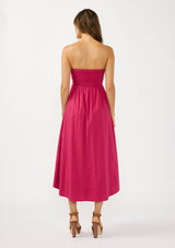 [Color: Magenta] A back facing image of a brunette model wearing a pink strapless midi dress with a gathered bust, side pockets, and a smocked back.