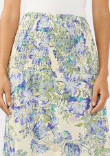 [Color: Cream/Dusty Lilac] A detail image of a brunette model wearing a cream and purple floral skirt with a smocked waist. Styled with a white bodysuit. 