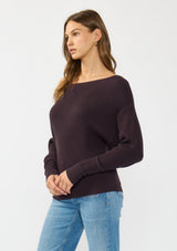[Color: Aubergine] A side facing image of a brunette model wearing a dark purple waffle knit pullover sweater. With long sleeves, a relaxed fit, and a wide neckline that can be worn off the shoulder.