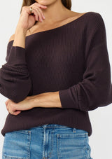 [Color: Aubergine] A detail image of a brunette model wearing a dark purple waffle knit pullover sweater. With long sleeves, a relaxed fit, and a wide neckline that can be worn off the shoulder.