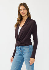 [Color: Aubergine] A side facing image of a Lovestitch model wearing a chic dark purple waffle knit wrap front sweater with long sleeves, a v neckline, and a button closure at the back. The long ties can be styled in multiple ways.