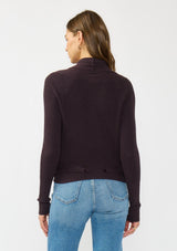 [Color: Aubergine] A back facing image of a Lovestitch model wearing a chic dark purple waffle knit wrap front sweater with long sleeves, a v neckline, and a button closure at the back. The long ties can be styled in multiple ways.