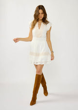 [Color: White] A front facing image of a blonde model wearing a boho white mini dress with delicate crochet lace accents. Featuring flirty short sleeves, a smocked waist, and a tiered ruffled hem. A daytime dress for casual and special occasions. 