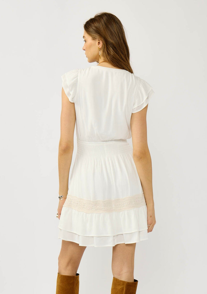 [Color: White] A back facing image of a blonde model wearing a boho white mini dress with delicate crochet lace accents. Featuring flirty short sleeves, a smocked waist, and a tiered ruffled hem. A daytime dress for casual and special occasions. 
