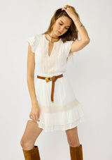 [Color: White] A front facing image of a blonde model wearing a boho white mini dress with delicate crochet lace accents. Featuring flirty short sleeves, a smocked waist, and a tiered ruffled hem. A daytime dress for casual and special occasions. 