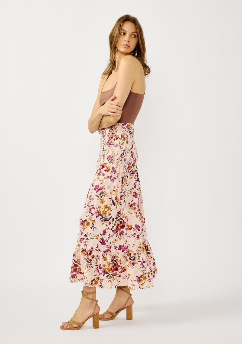 [Color: Almond/Wine] A side facing image of a blonde model wearing a convertible pink floral print skirt with a smocked waist and a ruffle trimmed hem. 