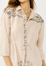 [Color: Natural/Cement] A detail image of a blonde model wearing a cotton gauze mini dress with embroidered details. Featuring a collared neckline, button front, a drawstring waist, and 3/4 length sleeves. 