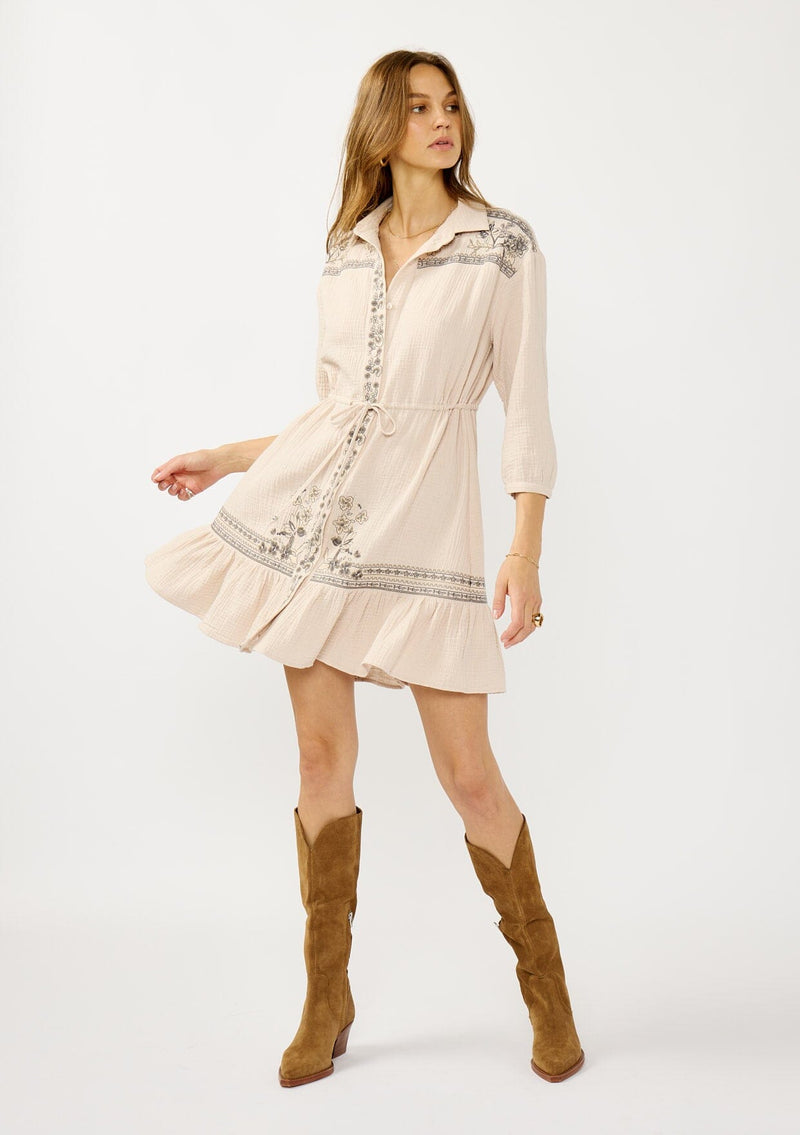 [Color: Natural/Cement] A front facing image of a blonde model wearing a cotton gauze mini dress with embroidered details. Featuring a collared neckline, button front, a drawstring waist, and 3/4 length sleeves. 