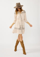 [Color: Natural/Cement] A front facing image of a blonde model wearing a cotton gauze mini dress with embroidered details. Featuring a collared neckline, button front, a drawstring waist, and 3/4 length sleeves. 