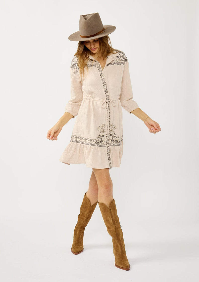 [Color: Natural/Cement] A front facing image of a blonde model wearing a cotton gauze mini dress with embroidered details. Featuring a collared neckline, button front, a drawstring waist, and 3/4 length sleeves. 
