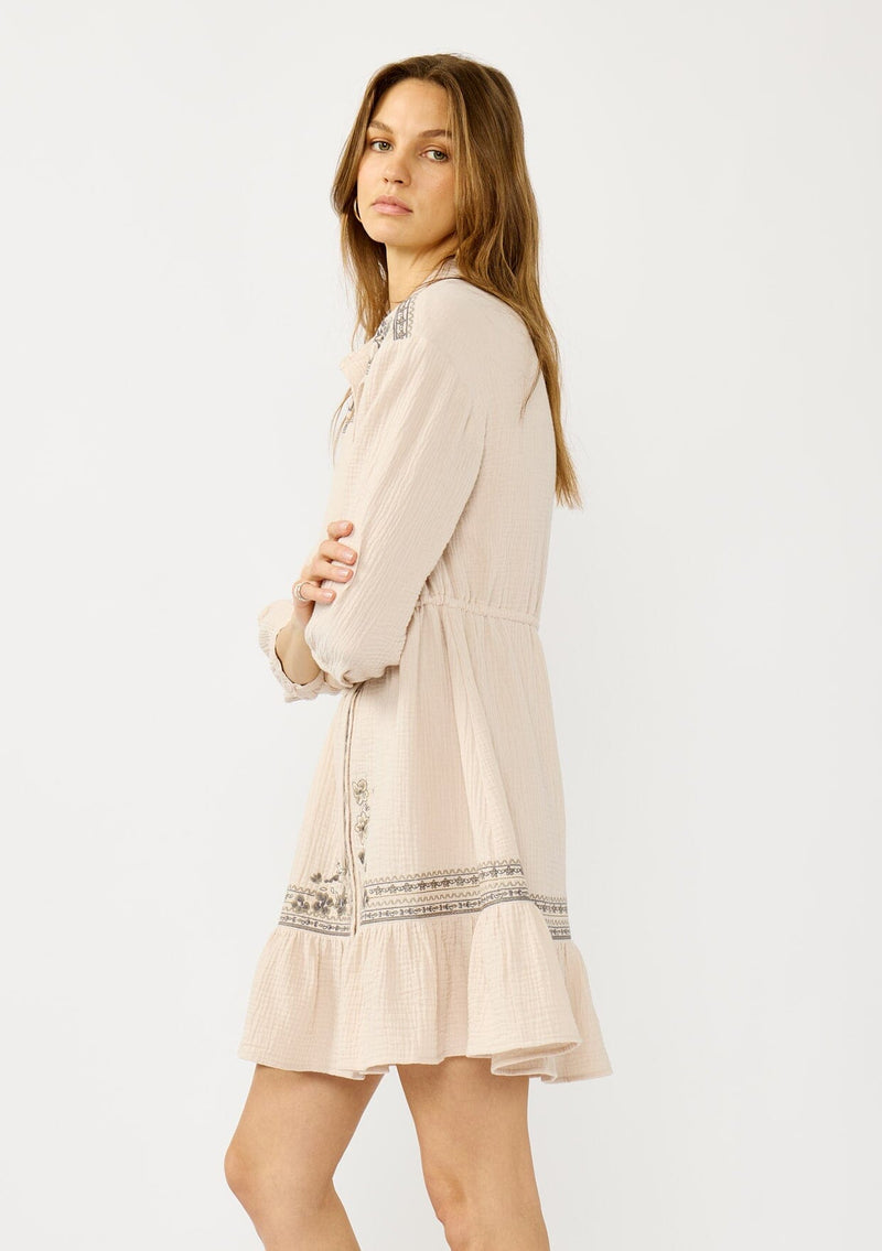 [Color: Natural/Cement] A side facing image of a blonde model wearing a cotton gauze mini dress with embroidered details. Featuring a collared neckline, button front, a drawstring waist, and 3/4 length sleeves. 