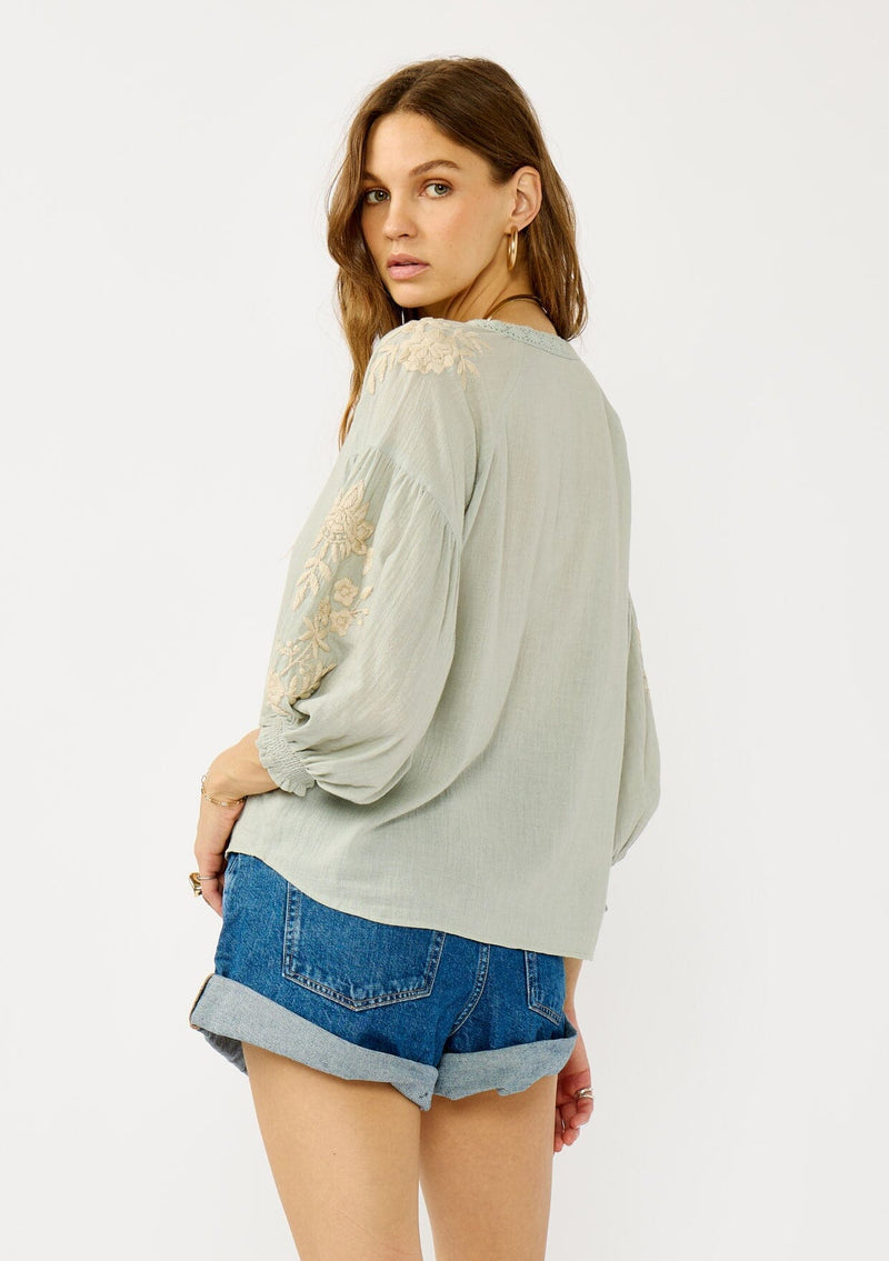 [Color: Dusty Sage/Natural] A side facing image of a blonde model wearing a green boho embroidered blouse with a split v-neckline, lace trim details, long balloon sleeves, and smocked cuffs. 