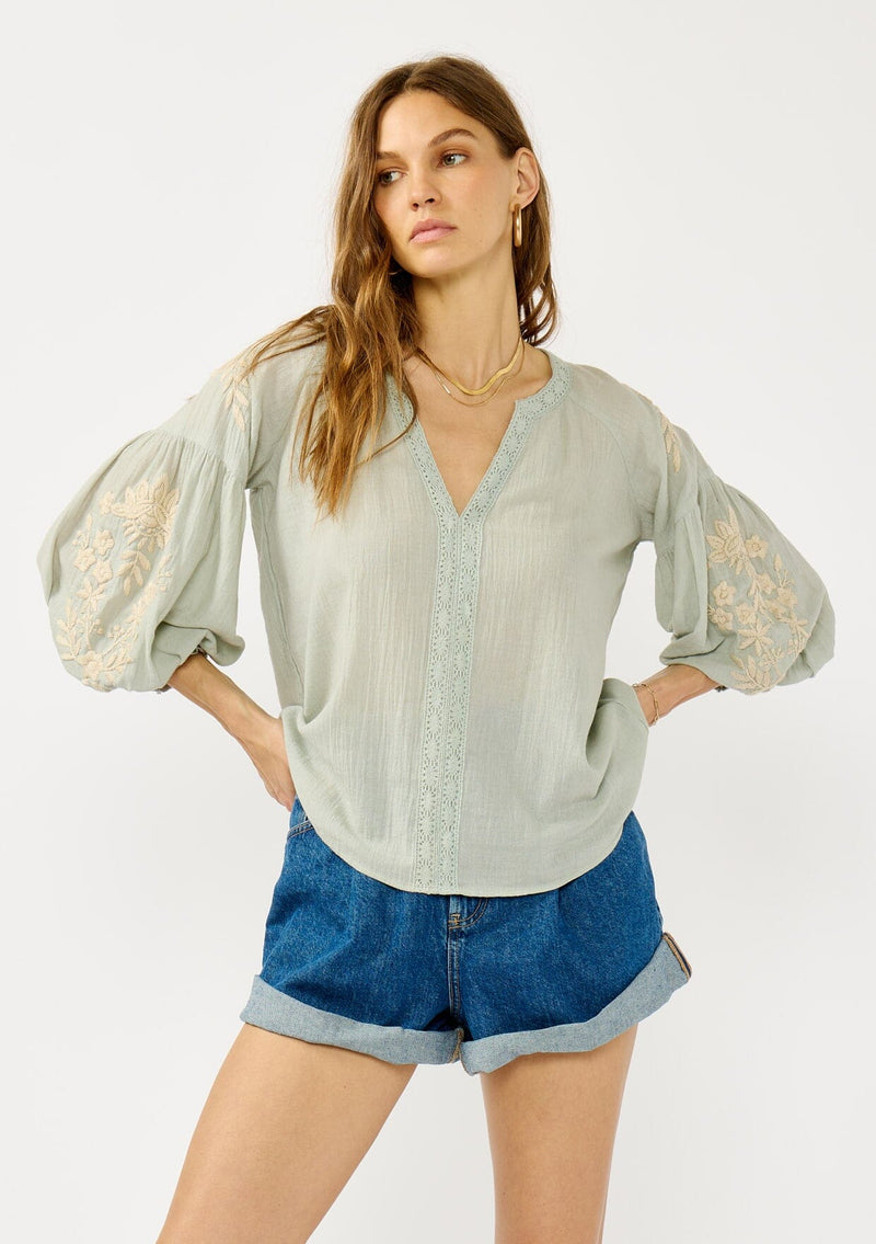 [Color: Dusty Sage/Natural] A front facing image of a blonde model wearing a green boho embroidered blouse with a split v-neckline, lace trim details, long balloon sleeves, and smocked cuffs. 