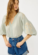 [Color: Dusty Sage/Natural] A front facing image of a blonde model wearing a green boho embroidered blouse with a split v-neckline, lace trim details, long balloon sleeves, and smocked cuffs. 