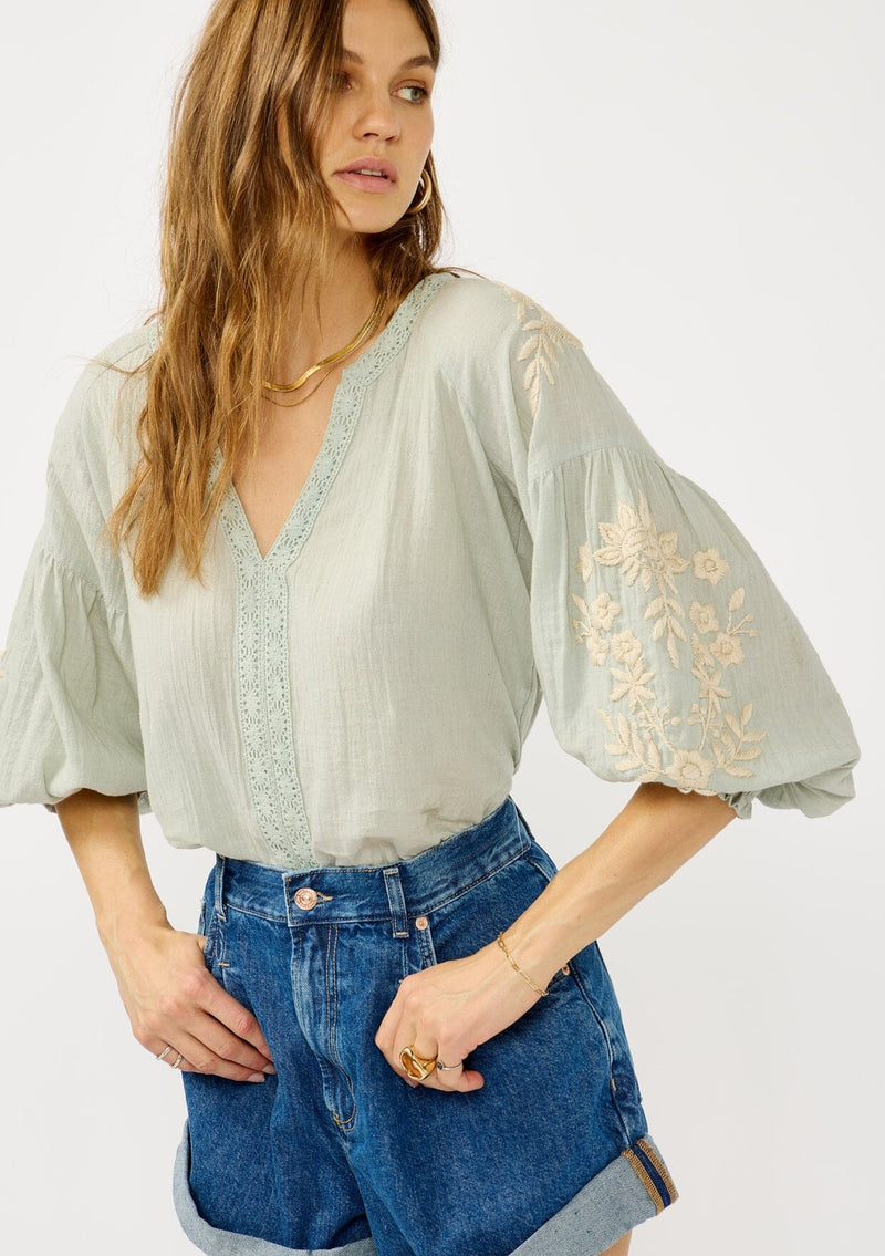 [Color: Dusty Sage/Natural] A front facing image of a blonde model wearing a green boho embroidered blouse with a split v-neckline, lace trim details, long balloon sleeves, and smocked cuffs. 