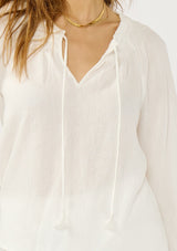 [Color: White] A detail image of a blonde model wearing an ultra boho relaxed white cotton blouse with a smocked split neckline, tie detail, and long sleeves with smocked cuffs. 