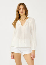 [Color: White] A front facing image of a blonde model wearing an ultra boho relaxed white cotton blouse with a smocked split neckline, tie detail, and long sleeves with smocked cuffs. 
