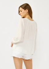 [Color: White] A back facing image of a blonde model wearing an ultra boho relaxed white cotton blouse with a smocked split neckline, tie detail, and long sleeves with smocked cuffs. 