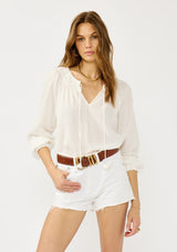 [Color: White] A front facing image of a blonde model wearing an ultra boho relaxed white cotton blouse with a smocked split neckline, tie detail, and long sleeves with smocked cuffs. 
