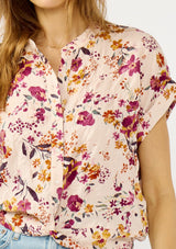 [Color: Almond/Wine] A detail image of a blonde model wearing a pink floral print top with a button front, twist front detail, and short dolman sleeves. 