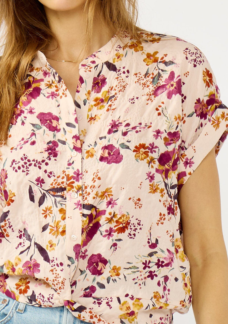 [Color: Almond/Wine] A detail image of a blonde model wearing a pink floral print top with a button front, twist front detail, and short dolman sleeves. 