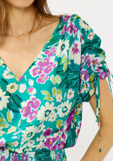 [Color: Teal/Plum] A detail image of a blonde model wearing a teal and purple floral print top with ruched dolman sleeves, a v-neckline, a smocked waistline, and a v-back with strap bridge. 