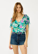[Color: Teal/Plum] A front facing image of a blonde model wearing a teal and purple floral print top with ruched dolman sleeves, a v-neckline, a smocked waistline, and a v-back with strap bridge. 