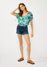 [Color: Teal/Plum] A front facing image of a blonde model wearing a teal and purple floral print top with ruched dolman sleeves, a v-neckline, a smocked waistline, and a v-back with strap bridge. 