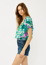 [Color: Teal/Plum] A side facing image of a blonde model wearing a teal and purple floral print top with ruched dolman sleeves, a v-neckline, a smocked waistline, and a v-back with strap bridge. 