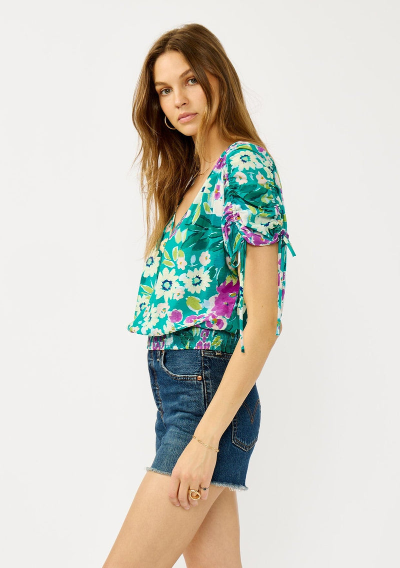 [Color: Teal/Plum] A side facing image of a blonde model wearing a teal and purple floral print top with ruched dolman sleeves, a v-neckline, a smocked waistline, and a v-back with strap bridge. 
