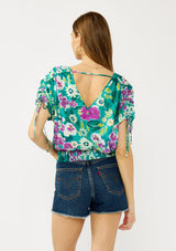 [Color: Teal/Plum] A back facing image of a blonde model wearing a teal and purple floral print top with ruched dolman sleeves, a v-neckline, a smocked waistline, and a v-back with strap bridge. 