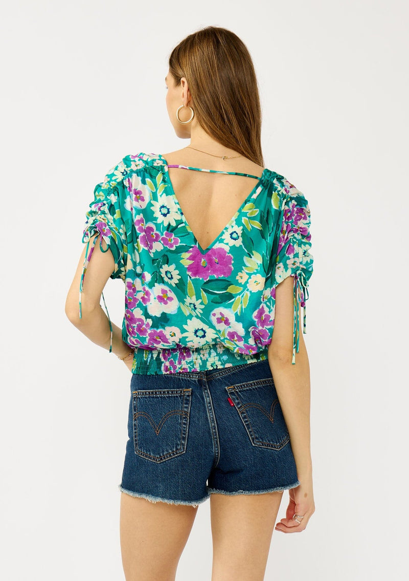 [Color: Teal/Plum] A back facing image of a blonde model wearing a teal and purple floral print top with ruched dolman sleeves, a v-neckline, a smocked waistline, and a v-back with strap bridge. 
