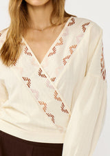 [Color: Vanilla/Rust] A detail image of a blonde model wearing an off white embroidered wrap top with dropped shoulders, long sleeves, and a side tie closure. An effortless boho top for the fall season. 