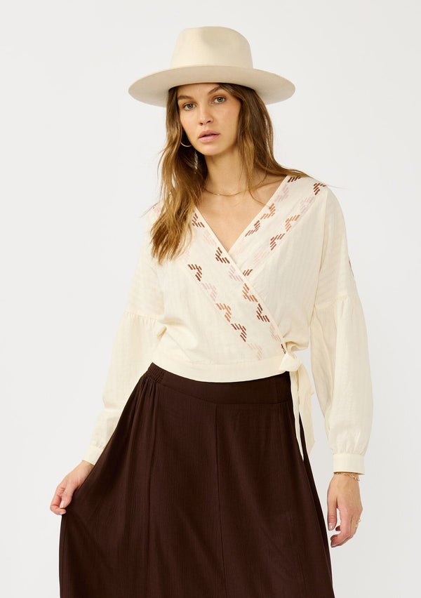 [Color: Vanilla/Rust] A front facing image of a blonde model wearing an off white embroidered wrap top with dropped shoulders, long sleeves, and a side tie closure. An effortless boho top for the fall season. 