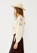 [Color: Vanilla/Rust] A side facing image of a blonde model wearing an off white embroidered wrap top with dropped shoulders, long sleeves, and a side tie closure. An effortless boho top for the fall season. 